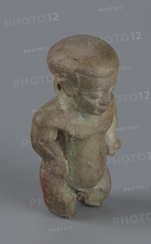Amulet - Ptah-dwarf Pataikos, Ptah-dwarf mythology. Figurine of a naked dwarf man with a large head with a wide forehead, short arms with clenched fists pressed to the hips, and short legs broken off at the knees. Careful modelling. 7th–4th century BC; Late Period, Egypt. Figurine, amulet. Height 6.5 cm; width 3.7 cm; depth 2.6 cm., A small sculpted figure standing on a plain gray background, with a rounded head and simplified facial features, a compact torso, and short limbs; the surface shows a weathered, mottled texture with shades of brown and green and hints of darker spots, and a small area of reddish coloration on one leg; the figure appears solid with smooth worn contours and subtle variations of pale greenish patina and brownish earth tones across its form.