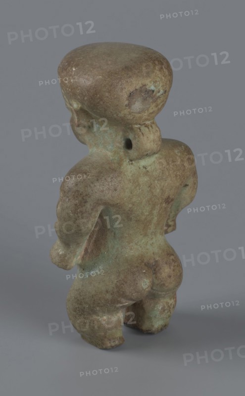 Amulet - Ptah-dwarf Pataikos, Ptah-dwarf mythology. Figurine of a naked dwarf man with a large head with a wide forehead, short arms with clenched fists pressed to the hips, and short legs broken off at the knees. Careful modelling. 7th–4th century BC; Late Period, Egypt. Figurine, amulet. Height 6.5 cm, width 3.7 cm, depth 2.6 cm., A small, weathered figurine shown from the back standing on a flat surface, with a rounded head, short neck, broad shoulders, and a rounded torso and legs; the surface shows an uneven, mottled texture with patches of greenish patina and areas of brown and tan discoloration, and a few darker spots; the background is a smooth, neutral gray.