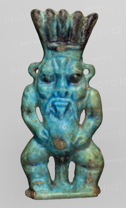 Amulet — god Bes; maker, Bes mythology, Egyptian deity; Late Period, 664–332 BC, Egypt; amulet; height 7.2 cm, width 3.2 cm., A small sculpted figure shown against a plain light gray background, rendered in shades of turquoise, blue, green, and patches of dark brown and tan; the figure has a flared, scalloped headdress that is darker at the top, a stylized face with deep-set eyes and a pronounced triangular beard area, and looped ear ornaments on each side; the torso is compact with arms bent at the elbows resting on the hips, and the lower body shows bent legs separated by two vertical openings, with worn spots and areas of surface discoloration in brown and tan.