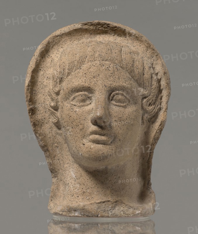 Votive head, Head of a young man covered with a shawl, from under which hair emerges in the form of a fringe and falls in curls below the temples, covering the ears. Large, wide-open, convex eyes; large, straight nose; narrow mouth with wide, slightly curled, pouting lips. Face with a strong chin alluding to classical traditions. 4th century BC, Central Italy; votive offering. Height 27.3 cm, width 20.5 cm, depth 18.5 cm, weight 3.5 kg., A weathered stone head sculpture shown against a plain gray background, featuring a rounded halo-like rim around the top and sides of the head, stylized short hair carved above the forehead, almond-shaped eyes without detailed pupils, a defined nose and closed lips, and carved ears partly visible beneath the hair; the surface shows a grainy texture with small dark speckles and areas of minor chipping and erosion. The visible colors are tan, beige, light brown, dark gray speckles, and a neutral gray background.