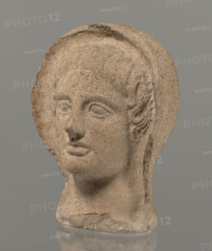 Votive head, Head of a young man covered with a shawl, from under which hair emerges in the form of a fringe and falls in curls below the temples, covering the ears. Large, wide-open, convex eyes; large, straight nose; narrow mouth with wide, slightly curled, as if pouting lips. The face with a strong chin alludes to classical traditions. 4th century BC, Central Italy, votive offering. Height 27.3 cm, width 20.5 cm, depth 18.5 cm, weight 3.5 kg., A carved stone head with a rounded top and a broken edge on one side sits against a plain gray background; the surface is sandy tan with darker speckling and small lighter flecks, showing carved facial features including shallow eye sockets, a defined nose with some surface damage at the tip, softly contoured lips, and a smooth neck that rests on a flat base; hair is indicated by shallow grooves and a few textured curls at the temple, and the stone shows areas of roughness and small pits throughout, with the whole object reflected faintly on t...