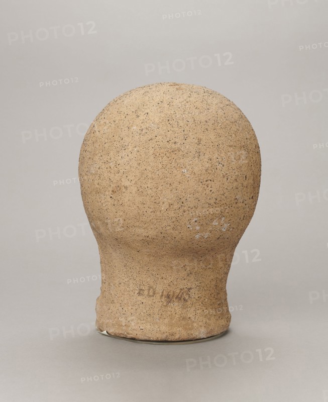 Votive head, Head of a young man covered with a shawl, from under which hair emerges in the form of a fringe and falls in curls below the temples, covering the ears. Large, wide-open, and convex eyes, large, straight nose, narrow mouth with wide, slightly curled, as if pouting lips. The face with a strong chin alludes to classical traditions. 4th century BC, Central Italy, votive offering. Height 27.3 cm, width 20.5 cm, depth 18.5 cm, weight 3.5 kg., A roughly spherical object with a narrower cylindrical base sits centered against a plain background, showing a coarse, textured surface that appears grainy and uneven; the object is primarily tan with darker brown speckles and small patches of white, while the background and surface it rests on are shades of light gray.
