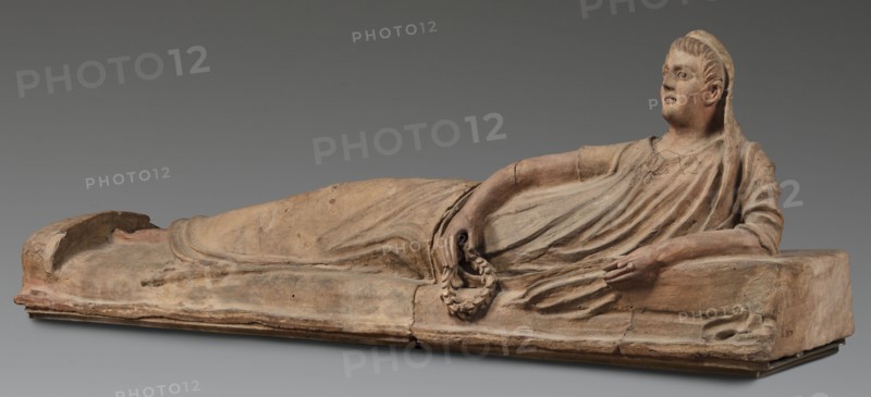 Sarcophagus lid, The deceased in a semi-reclining position, resting his left forearm on a pillow and holding a laurel wreath in his right hand. Dressed in a short-sleeved tunic and a cloak, which when thrown over the head reveals the torso and left arm from shoulder to elbow. The right foot is visible from under the cloak. Proportions of the human figure are very elongated, with especially long legs in disproportion to the torso. 2nd half of the 2nd century BC, Etruria. Sarcophagus; height 71 cm, width 203 cm, depth 48 cm, weight 108 kg., A reclining terracotta figure rendered in shades of beige, tan, brown, and hints of ochre rests on a rectangular base of similar earthy colors, showing the drape of a long garment with flowing folds across the body and arm. The figure lies on its side with a head turned upward, short stylized hair, and a cloth or veil falling behind it; a relaxed arm supports the upper body while the other hand holds a circular wreath-like object. The surface shows we...