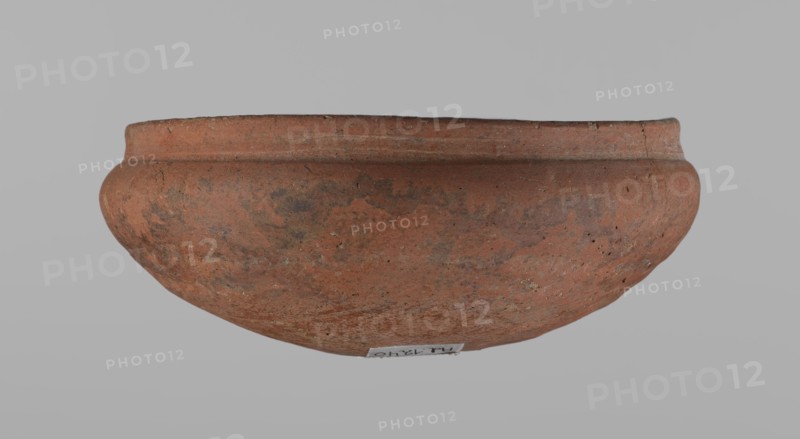 Medum-type bowl, Ceramics: Egyptian pottery vessel, clay bowl with hemispherical shape. The walls bend gently around about 2/3 of the height; rim straight and perpendicular to the shoulders, slightly irregular; rounded bottom. Surface even; vessel color red. Assembled from several fragments. c. 2686–c. 2181 BC Old Kingdom, from Edfu. Vessel; height 5.5 cm, diameter 12.6 cm rim, diameter 13.5 cm shoulders., A broad, shallow clay vessel with a rounded body and a slightly flared rim sits against a plain light gray background, showing a rough, weathered surface with mottled reddish-brown and darker brown areas and some small pits and specks of lighter beige.