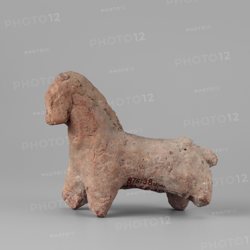 Horse figurine, horses, 664–30 BC; Late Period–Ptolemaic Period, found at Edfu, figurine, height 8 cm, width 10 cm, A small rough-textured sculpted figure of an animal with a rounded head, short neck, sturdy body, and four thick legs, shown in profile on a flat surface; the figure's surface has uneven, pitted areas and visible cracks. Colors visible include shades of reddish-brown, terracotta, light brown, and small patches of pale gray, set against a neutral light gray background.