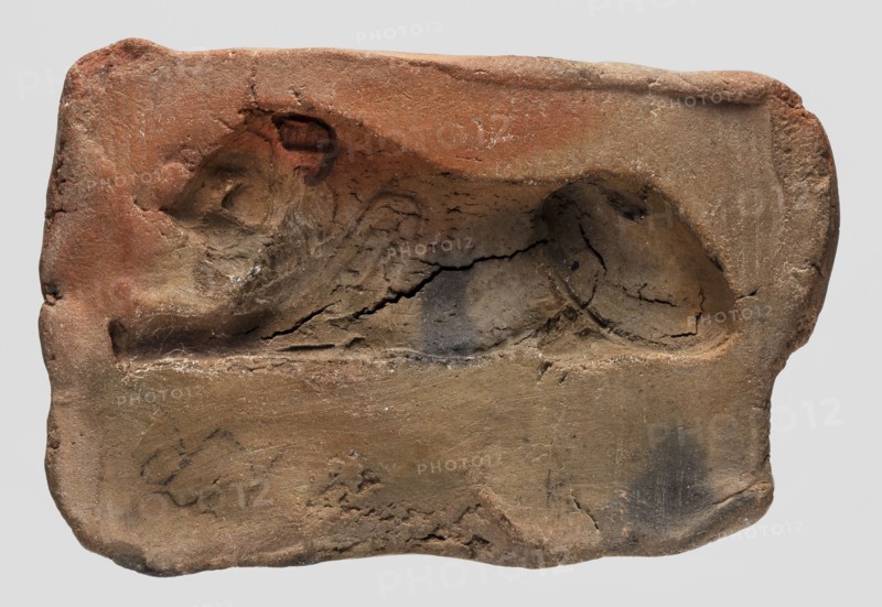 A mold for making an amulet lion, lions, casting mold, Ptolemaic Period 332–30 BC, found in Edfu; mold; height 7.1 cm., A rectangular, worn clay object in shades of reddish-brown, tan, beige, and dark gray with an indented relief showing a reclining feline-like form; the surface is cracked and pitted with rough, uneven edges and several small chips, and there are darker smudges and patches across the lower and central areas.