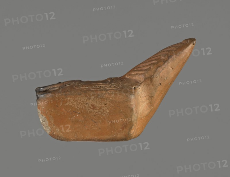 Oil lamp, unknown Pannonian workshop. Oil lamp with leaf ornament, Roman three-burner lamp. Made from a mold. Rectangular, flat vessel with a slightly recessed rectangular disc surrounded by a stylized egg-shaped ornament. Three olive holes placed at regular intervals within the disc. Leaf-shaped handle on the longer side. Opposite side has three short circular burners with large wick holes; all are charred. 1st–2nd century, Pannonia. Oil lamp; height 3.3 cm, width 7.3 cm, depth 5.1 cm., A single clay object photographed against a plain gray background, showing a worn rounded base and an elongated pointed extension; the surface is textured with irregularities and shallow decorative impressions, and there are rough patches and small pitted areas. The visible colors include shades of brown, orange-brown, and tan, along with some darker brown and grayish spots.