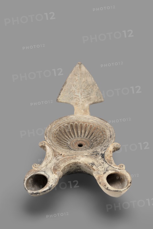 Two-burner lamp decorated with a rosette motif, oil lamp, end of the 1st century, Asia Minor; height 10.2 cm, width 21.5 cm, depth 13.5 cm, weight 0.260 kg., An off-white and beige ceramic object with areas of darker gray and black discoloration, shaped with a central shallow bowl featuring radiating fluted ridges and a small central hole, a triangular vertical backplate rising from the rear with faint carved floral-like motif, and two rounded protruding spouts at the front connected by a curved bridge, all resting against a plain mid-gray background.
