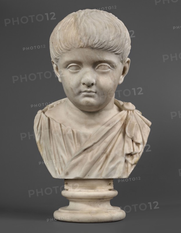 Portrait of a prince from the Julio-Claudian dynasty, unknown Roman workshop, boy portrait, Roman sculpture, antique bust, dated 40–70 CE, Roman Empire 27 BC–476, sculpture; height 36 cm, width 21 cm, depth 18 cm, weight 11.55 kg, A marble bust of a young child is shown against a gray background, carved with short textured hair and smooth facial features including closed or blanked eyes, a rounded nose, and a small mouth; the child wears a draped garment with folds and a shoulder knot, and the sculpture rests on a rounded cylindrical base. The visible colors are white and off-white tones in the marble with subtle beige and pale yellow discolorations, and a solid mid-gray background.