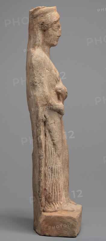 Figurine of a woman standing on a rectangular base. The woman is dressed in a finely pleated chiton reaching down to her bare feet, with a semicircular neckline. A himation, draped over her shoulders like a shawl, covers her arms to the elbows. On her head she wears a cylindrical polos, slightly widening upwards and folded into a veil flowing down the back. Hair protrudes from beneath the headdress, flowing in wavy patterns to the sides. The face is full, with a rounded chin, large eyes, and fleshy lips drawn into a characteristic smile. In her left hand, bent at the elbow, the woman holds a piglet; with her right hand she supports the animal's hind leg. Pigs iconography; female figurines. 5th century BC, Southern Italy Italy. Terracotta figurine; height 28 cm, width 10 cm, depth 10 cm, weight 0.57 kg., A weathered stone statue of a standing female figure shown in profile, with a tall headdress or hair pulled back, a visible nose and chin, and a calm facial expression; the figure's arm...