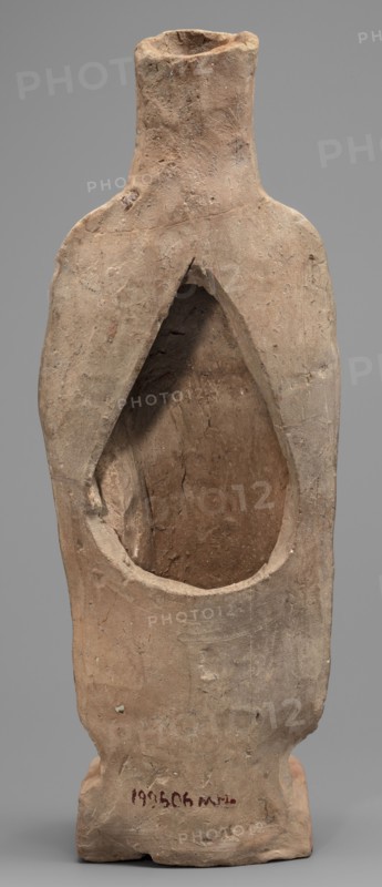 Figurine of a woman standing on a rectangular base. The woman is dressed in a finely pleated chiton reaching down to her bare feet, with a semicircular neckline. A himation, draped over her shoulders like a shawl, covers her arms to the elbows. On her head she wears a cylindrical polos, slightly widening upwards and folded into a veil flowing down the back. Hair protrudes from beneath the headdress, flowing in wavy patterns to the sides. The face is full, with a rounded chin, large eyes, and fleshy lips drawn into a characteristic smile. In her left hand, bent at the elbow, the woman holds a piglet; with her right hand she supports the animal's hind leg. 5th century BC, Southern Italy Italy. Terracotta figurine, height 28 cm, width 10 cm, depth 10 cm, weight 0.57 kg. Iconography: pigs; category: female figurines., A roughly textured hollow clay object shaped like a stylized standing figure with a narrow neck and flared shoulders, showing an open teardrop-shaped cavity in its front; the...