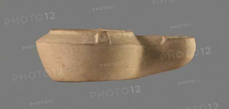 Lamp with comic mask, unknown Italian workshop. Roman oil lamp, Buchi type IX-b = Loeschcke type IXb, from a mould. Round vessel with arms drooping to the outer, separated from the disc by a convex edge; two bars arranged symmetrically on the arms. Concave, flat disc with one olive hole located below a convex theatrical comic mask. Medium-length wick channel decorated with a central groove; rounded burner end. No traces of use no charring. Convex inscription workshop trademark on the bottom: STROBILI. 1st century AD Flavian period, 2nd half of the 18th century, Italy — Magreta, near Modena; Modena. Oil lamp, height 3.4 cm, width 10.1 cm., A single, beige clay object with a smooth, rounded form is shown against a solid gray background; the object has a shallow, open top and an irregular rim with a few raised sections and indentations, a visibly textured surface with subtle horizontal tool marks and small pits, and areas of slightly darker beige shading where the surface curves.