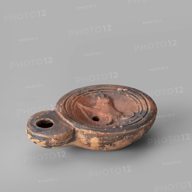 Lamp decorated with a depiction of a dog, unknown eastern workshop, single-burner oil lamp, 1st century, Roman Empire 27 BC–476, weight 0.082 kg, dogs iconography., The image shows an oval-shaped terracotta oil lamp with a rounded handle extension, placed on a smooth gray surface and set against a gray background. The lamp has a central shallow bowl with a small round hole and a molded raised motif in the center, surrounded by a series of concentric ridges. The handle has a circular opening. The surface of the lamp shows worn and rough textures with patches of darker discoloration and small chips. Visible colors include reddish-brown, dark brown, blackened areas, and the surrounding gray background and surface.