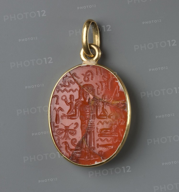 Medallion with a forged gem, France? Italy?, forgery of an oval gem set in a gold frame with an oval pendant. The gem's surface bears a carved image of a standing woman in a long robe with her arms bent at right angles in a gesture of prayer?. She wears a wig with curls falling over her shoulders. Various pseudo-hieroglyphs are carved around the figure. 17th–18th century; modern period, jewelry, weight 6.42 g, An oval pendant with a golden metal frame and loop, containing a polished reddish-orange stone carved with a central standing human figure and a surrounding array of abstract and decorative carved motifs; the stone shows a few visible cracks and fissures, and the background behind the pendant is a uniform medium gray.