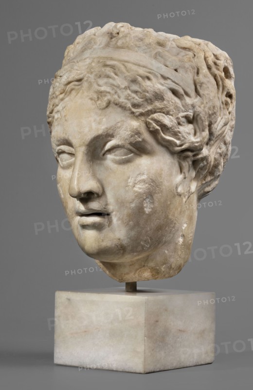 Head of Venus or a nymph, unknown Roman workshop, female portrait, Roman sculpture, hairstyles, Roman copy after a Greek original from the 3rd century BC, early 1st century period of Octavian Augustus, Roman Empire, 27 BC–476, sculpture; height 23 cm, width 16 cm, depth 19 cm, weight 11.13 kg., A sculpted head displayed on a rectangular block, carved from a pale stone with areas of weathering and discoloration, depicting a face with closed or partially closed eyes, a straight nose, and slightly parted lips; the hair is rendered in textured, wavy locks gathered back and partially covered by a band, and the surface shows patches of roughness and subtle variations in beige, off-white, cream, light brown, and gray tones against a uniform gray background.