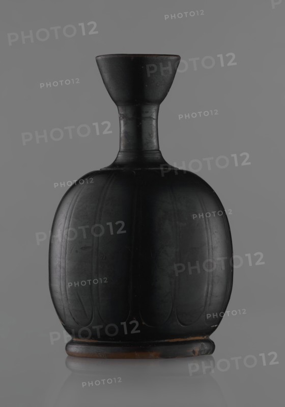 Black-finished aryballoi lekythos, Greek black-glazed pottery. Aryballoi lekythos coated with black varnish. Medium with flat foot flowing directly into a bulging body, significantly widened in its lower section. Nearly horizontal handles flow into a slender neck ending in a markedly widening, cup-shaped rim. Handle attached at the transition from body to handles and below the cup-shaped rim. Body bears an engraved ornament of long, vertically arranged petals. Good condition, minor damage to varnish; neck cracked and glued. 4th century BC, Attic manufacture, vessel lekythos., A dark rounded vase with a narrow neck and flared rim set against a gray background, the vase appears primarily black with subtle brown tones at the rim and base, the body shows vertical carved or molded panels creating a ribbed appearance, the surface has a matte finish with faint lighter scuffs and worn spots, and the base sits on a slightly raised foot that shows more of the brown underlayer.