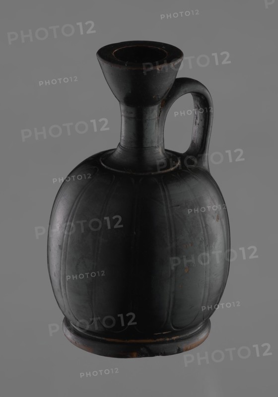 Black-finished aryballoi lekythos, Greek black-glaze pottery. Aryballoi lekythos coated with black varnish. The medium, flat foot flows directly into the bulging body, which is significantly widened in its lower section. The nearly horizontal handles flow into a slender neck ending in a significantly widening, cup-shaped rim. The handle is attached at the transition from the body to the handles and below the cup-shaped rim. The body bears an engraved ornament of long, vertically arranged petals. Good condition, with minor varnish loss; the neck is cracked and glued. 4th century BC, Attica, vessel, lekythos., A dark rounded ceramic jug with a bulbous body and a narrow neck leading to a flared mouth, featuring an arched handle attached between the neck and upper body; the surface is smooth with faint vertical ridges and small worn spots, and the visible colors are predominantly black with areas of dark brown and small touches of reddish-brown against a muted gray background.