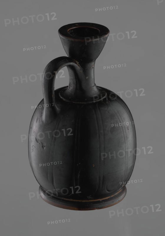 Black-finished aryballoid lekythos coated with black varnish. The medium, flat foot flows directly into the bulging body, which is significantly widened in its lower section. The nearly horizontal arms flow into a slender neck ending in a significantly widening, cup-shaped rim. The handle is attached at the transition from the body to the arms and below the cup-shaped rim. The body bears an engraved ornament of long, vertically arranged petals. Good condition, with minor damage to the varnish; the neck is cracked and glued. 4th century BC, Attic, Greek pottery — vessel lekythos., A single dark vessel with a rounded body and a narrow neck topped by a flared rim, shown against a plain gray background; the vessel has a curved handle connecting the neck to the shoulder and vertical rib-like grooves on its body; the surface shows small scuffs and worn spots revealing a reddish-brown underlayer beneath the dominant black finish; visible colors include black, gray, and reddish-brown.