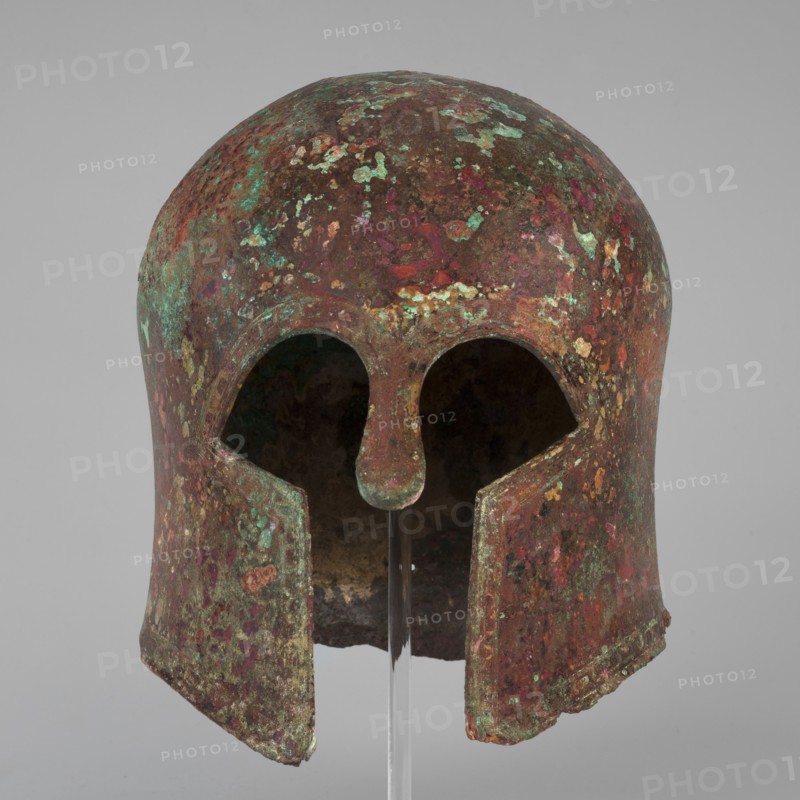 Corinthian-type helmet, This Corinthian-type helmet, with thicker walls in the cheekpieces and a significantly thickened nose plate, has a domed peak, beneath which the walls are slightly concave, widening again at the edge. At the back, near the edge, are two small, round holes for attaching a cord. There is a similar hole in the peak. The edges of the helmet, cheekpieces, and eyeholes are decorated with a precisely crafted band of ornamentation consisting of rectangular fields framed at the top and bottom by a double engraved line and separated by double engraved squares, between which are rows of tiny plastic spheres. A row of similar spheres is located between the lines framing the rectangles at the top. The ornamentation reaches the base of the nose guard, which is decorated only with engraved lines, repeating the shape of the protective plate. Dated c. 600 BC formerly 630–600 BC. Possible origin: Sparta Greece or Mycenae Greece according to Otto Schaubert. Function: weapon/helmet...