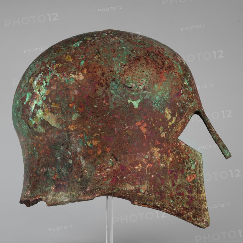 Corinthian-type helmet. This helmet, with thicker walls in the cheekpieces and a significantly thickened nose plate, has a domed peak beneath which the walls are slightly concave, widening again at the edge. At the back, near the edge, are two small round holes for attaching a cord; there is a similar hole in the peak. The edges of the helmet, cheekpieces, and eyeholes are decorated with a precisely crafted band of ornamentation consisting of rectangular fields framed at the top and bottom by a double engraved line and separated by double engraved squares, between which are rows of tiny applied spheres. A row of similar spheres lies between the lines framing the rectangles at the top. The ornamentation reaches the base of the nose guard, which is decorated only with engraved lines repeating the shape of the protective plate. Dated ca. 600 BC previously dated 630–600 BC. Possible origin: Sparta or Mycenae according to Otto Schaubert. Function: weapon. Height 23 cm; width 18.2 cm; depth ...