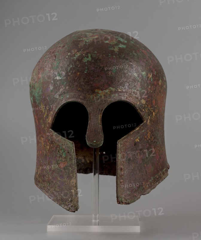 Corinthian-type helmet, This Corinthian-type helmet, with thicker walls in the cheekpieces and a significantly thickened nose plate, has a domed peak, beneath which the walls are slightly concave, widening again at the edge. At the back, near the edge, are two small, round holes for attaching a cord. There is a similar hole in the peak. The edges of the helmet, cheekpieces, and eyeholes are decorated with a precisely crafted band of ornamentation consisting of rectangular fields framed at the top and bottom by a double engraved line and separated by double engraved squares, between which are rows of tiny plastic spheres. A row of similar spheres is located between the lines framing the rectangles at the top. The ornamentation reaches the base of the nose guard, which is decorated only with engraved lines, repeating the shape of the protective plate. Date: 600 BC previously dated 630–600 BC. Place of origin: Sparta Greece, — Mycenae Greece ? according to Otto Schaubert. Function: weapon...