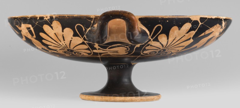 Kylix depicting youths at play, Thalia Painter fl. 520–510 BC. A: three naked youths; B: two youths with spears, one with a rhyton. Palmettes at the handles, lotus buds under the handles. At the bottom, a reclining naked youth. Sport in antiquity iconography. Greek ceramics; vessel kylix. 510–500 BC previously dated c. 520 BC. Attica. Weight 1.030 kg., A shallow pedestal bowl with a curved handle rising from its rim, decorated with a floral motif in contrasting colors; the bowl and pedestal are primarily black with beige or tan painted designs and areas of wear, showing rougher beige edges and cracks; the floral elements include petal-like shapes and spiral forms in beige against the black background, and the handle shows both black and beige tones where the surface is worn.