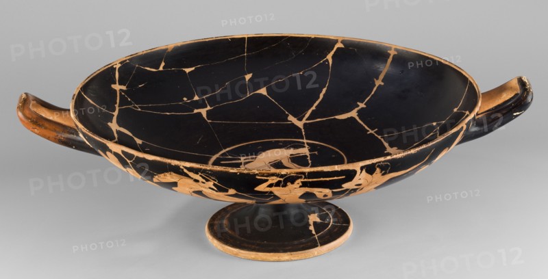 Kylix depicting youths at play, Thalia Painter fl. 520–510 BC; scene: A: three naked youths; B: two youths with spears, one holding a rhyton; at the bottom, a leaning naked youth. Decoration includes palmettes at the vices and lotus buds under the vices. Greek ceramic vessel kylix, Attica; dated 510–500 BC previous dating c. 520 BC. Weight 1.030 kg., A shallow, wide bowl with two horizontal handles sits on a low pedestal; the bowl's interior and exterior surfaces show a dark, almost black finish interrupted by a network of lighter, tan-colored cracks and repaired lines, and the rim, handles, and foot display exposed tan areas where the dark surface is worn. The handles are elongated and curved with dark outer surfaces and tan-colored inner faces, and the pedestal foot is dark with tan edges and a visible patch of lighter tone. The interior shows a smooth dark field with irregular tan fissures, while the exterior includes silhouettes and partial figures in the tan color against the dark...