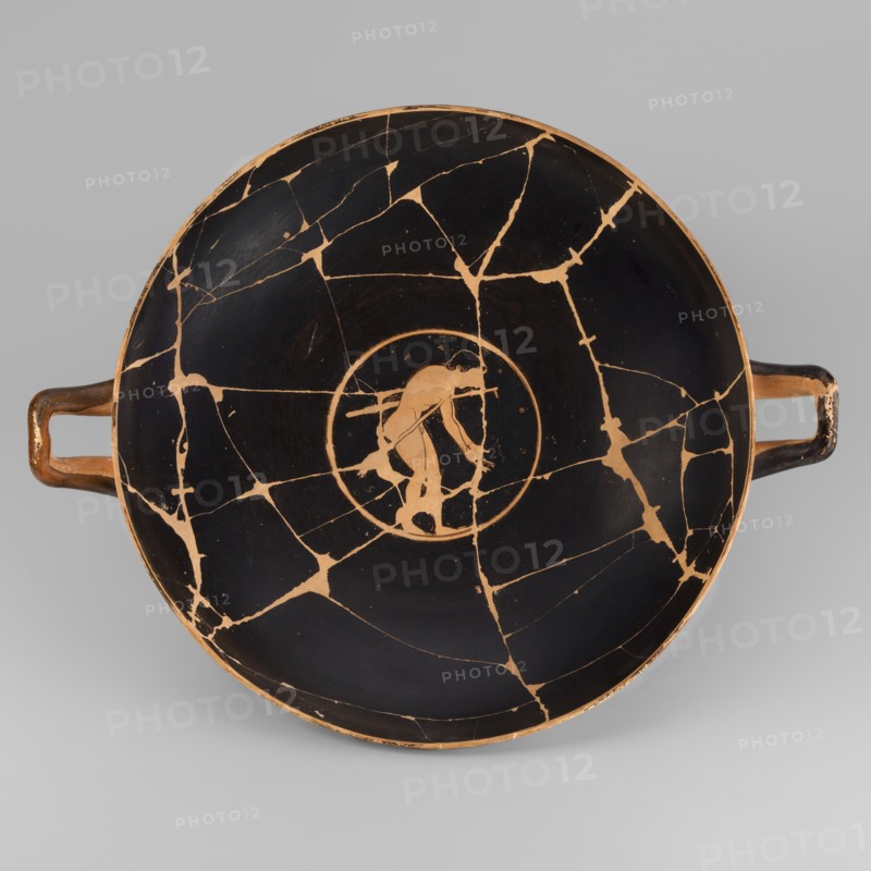 Kylix depicting youths at play, Thalia Painter fl. 520–510 BC. A: three naked youths; B: two youths with spears, one with a rhyton. Palmettes at the handles, lotus buds under the handles. At the bottom, a leaning naked youth. Sport in antiquity iconography; Greek ceramics; vessel kylix. 510–500 BC formerly dated c. 520 BC. Attica. Weight 1.030 kg., A shallow round bowl with two horizontal handles, shown from above, with a black interior and orange-brown lines and areas where the black surface is broken into a network of cracks; at the center a circular orange-brown medallion contains the silhouette of a person in orange-brown against the black, and the handles are orange-brown with worn black areas.