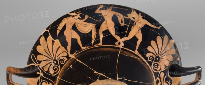 Kylix depicting youths at play, Thalia Painter 520–510 BC. A: three naked youths; B: two youths with spears, one with a rhyton. Palmettes at the vices, lotus buds under the vices. At the bottom, a leaning, naked youth. Sport in antiquity iconography. Greek ceramics; Attica; vessel kylix. Weight 1.030 kg. Date: 510–500 BC previous dating c. 520 BC. Thalia Painter fl. 520–510 BC., A shallow curved ceramic vessel fragment with a black background and orange figures and decorative motifs, showing a partial scene of three nude human figures in orange with black detailing positioned along the rim, each in different dynamic poses and one holding a curved object, flanked by large stylized orange palmette motifs with swirling elements on either side; the surface shows visible cracks and areas of wear where the orange slip is chipped, and the vessel has two short black handles extending outward from the lower edges.