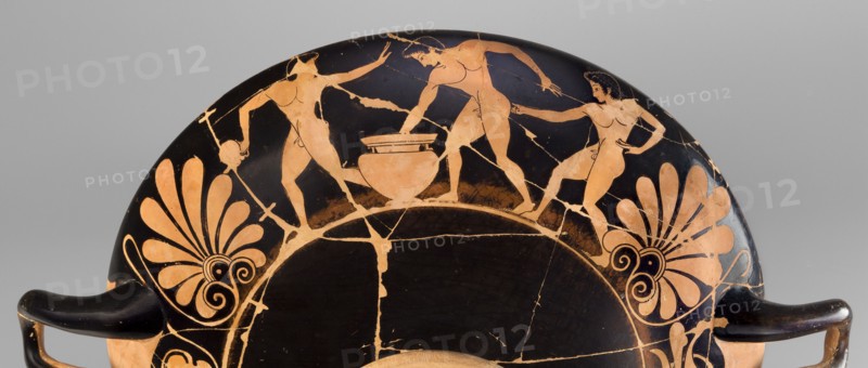Kylix depicting youths at play, Thalia Painter 520–510 BC. Red-figure kylix, Attica, dated 510–500 BC formerly c. 520 BC. Interior A: three naked youths; exterior B: two youths with spears, one with a rhyton. Palmettes at the handles, lotus buds under the handles; at the bottom, a leaning naked youth. Subject: sport in antiquity. Maker: Thalia Painter fl. 520–510 BC. Vessel, weight 1.030 kg., A fragmentary round ceramic vessel rim with black and terracotta colors shows a painted scene of three nude human figures around a central vessel, with two standing figures reaching toward it and one figure holding a staff; decorative palmette motifs in terracotta appear on either side against the black background, and the piece displays visible craquelure and repaired breaks, with the vessel handles partially visible at the lower edges in black.