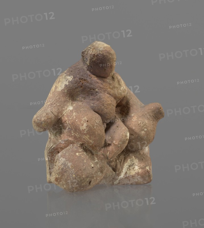 Terracotta figurine of Silenus, Silenus mythology; terracotta. Figurine of an obese Silenus sitting on a rock. The figurine is hollow inside, open at the bottom. The back is only roughly worked, with a large, triangular opening. The clay is red, strongly and unevenly fired; the surface is light, cracked, and mostly chipped. The idol sits frontally, with thighs widely spread and feet crossed. The belly is sagging. The right arm rests with its elbow on the thigh, the forearm and hand stretched slightly downward. The left hand holds a waterskin resting on the rock and the side of the thigh. The small head, slightly lowered, turns to the left and downward, towards the waterskin. Silenus is partially covered by a chlamys, fastened at the right shoulder and thrown back over the left arm; the lower torso and legs are bare. Schematic modeling; head, arms, legs and waterskin rendered synthetically, with a tendency to accentuate volume. Numerous chips and surface flaking. Wide crack in the base ...