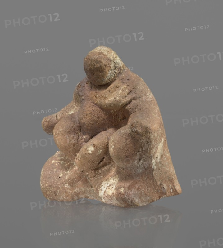Terracotta figurine of Silenus, Silenus mythology, terracotta. Figurine of an obese Silenus sitting on a rock. The figurine is hollow inside, open at the bottom. The back is only roughly worked, with a large triangular opening. The clay is red, strongly and unevenly fired; the surface is light, cracked, and mostly chipped. The idol sits frontally, thighs widely spread and feet crossed. The belly is sagging. The right arm rests with its elbow on the thigh, the forearm and hand stretched slightly downward. The left hand holds a waterskin resting on the rock and the side of the thigh. The small head, slightly lowered, turns to the left and downward, toward the waterskin. Silenus is partially covered by a chlamys, fastened at the right shoulder and thrown back over the left arm; the lower torso and legs are bare. Schematic modeling; head, arms, legs and waterskin rendered synthetically, with a tendency to accentuate volume. Numerous chips and surface flaking. Wide crack in the base on the ...