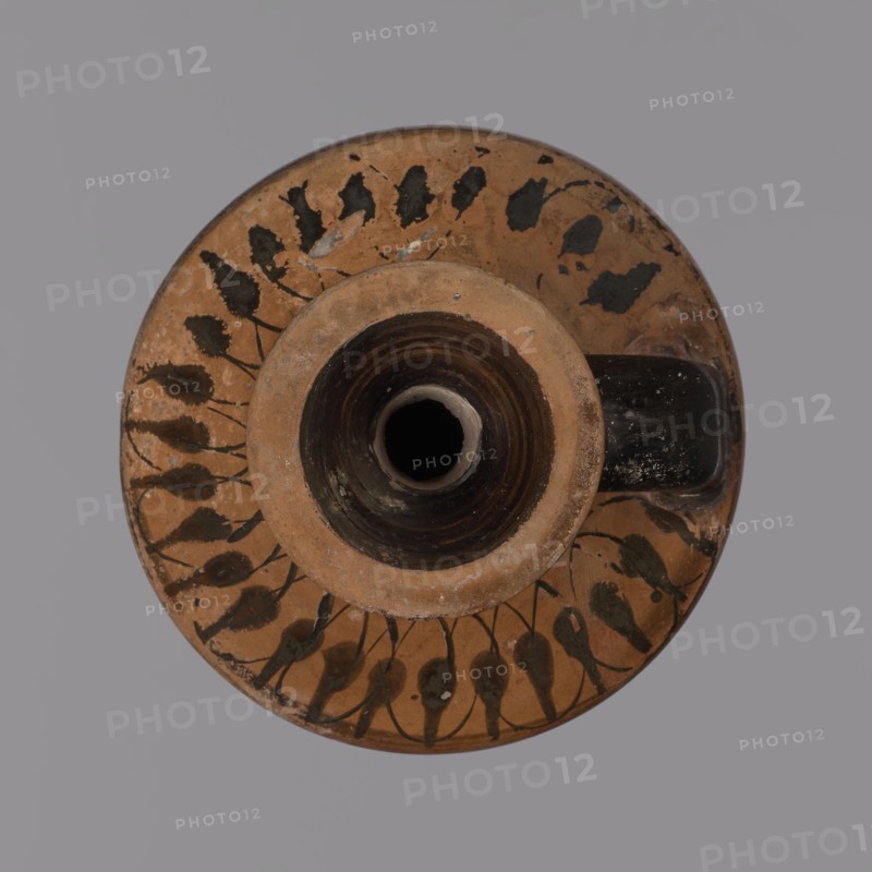 Black-figure lekythos, unknown Attic workshop. Yellowish clay, matte firnia. Disc-shaped foot, ovoid body, slightly sloping handles, wide short neck, rim widening upward. Dark firnia on the rim, outer part of the neck, lower body, and foot. A wide black band below the figural decoration and a narrow black band at the transition from body to handles. Handles decorated with a series of lotus buds connected by intertwining lines; neck with a circular ornament of dots. Belly figural scene: a maenad in a chiton and cloak dancing, facing right with her head turned back; her left arm raised upward and her right bent arm hidden under her cloak. Two dancing satyrs flank her, facing right. Vine branches with grapes in the background. Black-figure pottery, lekythos, c. 500 BC, Attica., A round ceramic object viewed from above with a central cylindrical opening surrounded by a raised rim, showing a terracotta base color with dark brown to black painted leaf-like motifs arranged in a circular patte...
