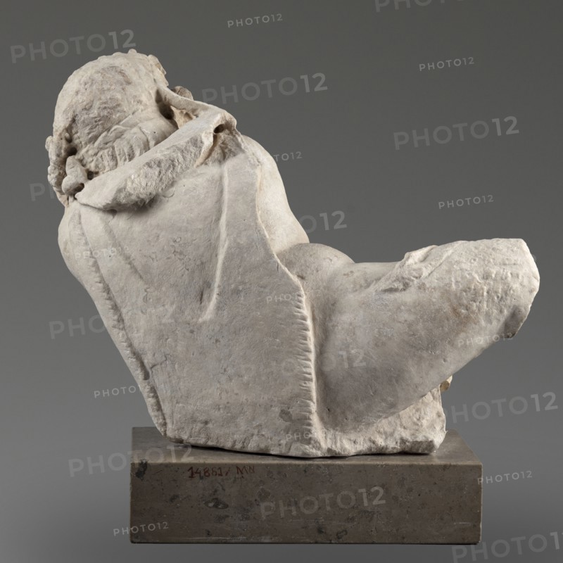 Reclining Silenus, unknown Roman workshop, Scen.Gal.Class., architectural elements, Silenus mythology, Roman sculpture — Sculpture of a reclining Silenus supported on his left hip and elbow, dated 69–79, Roman Empire 27 BC–476, sculpture; height 29.4 cm, width 37 cm, depth 22 cm, weight 19.4 kg, A weathered pale stone sculpture of a reclining figure is shown from a rear-quarter angle, resting on a rectangular darker stone base; the sculpture displays textured carved hair and draped cloth with visible folds and ridges, and a rounded limb extending outward; the colors visible are shades of off-white and light gray on the sculpture, darker gray on the base, and a neutral medium gray background.