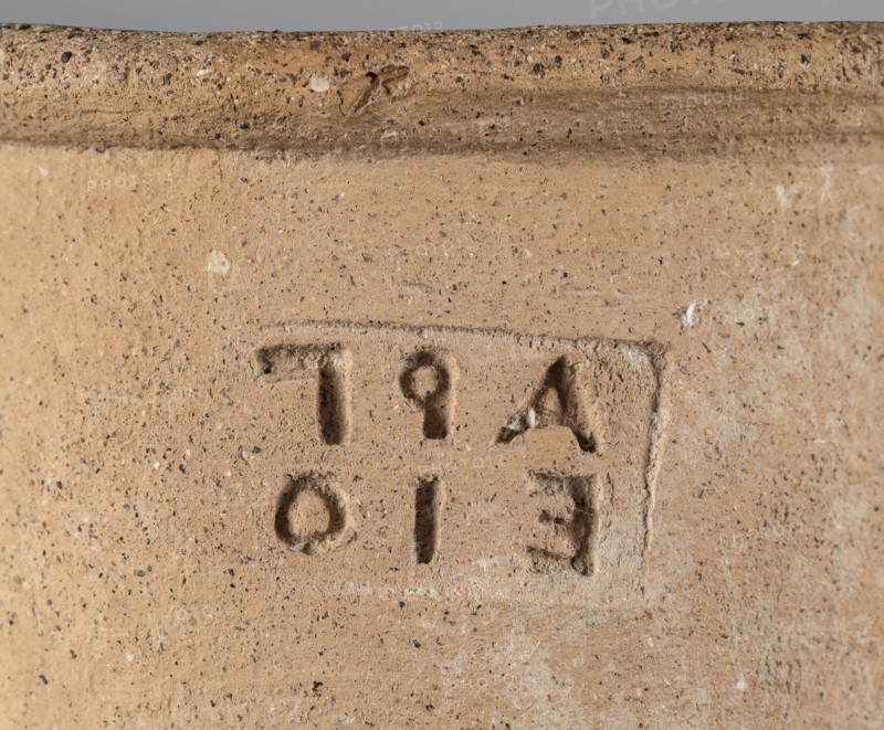 Stamped transport amphora with a Greek inscription Argeio — transport amphora with a pointed bottom and a conical belly, rounded at the sloping shoulders, merging into a cylindrical long neck slightly bulging under the annular rim. The neck is stamped; letters arranged in two rows. Ribbon-like handles protrude from the shoulders and run straight up, bending in an arc where they join the neck under the spout. A rectangular stamp is impressed on the upper part of the neck; the deeply recessed letters are arranged in two rows and refer to the maker or an official. Dated 400–390 BC. From Heraclea Pontica Bithynia; found at Olbia. Vessel type: transport amphora. Weight 6.326 kg; height 63 cm; diameter 26.5 cm., A close-up of a textured surface that appears to be a piece of terracotta or clay in warm beige and tan tones with small dark flecks throughout, showing an impression or recessed rectangular area with carved shapes and rounded depressions, and a raised rim along the top edge in sligh...
