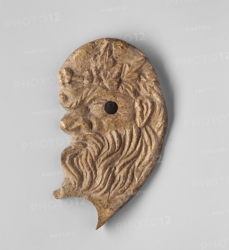 Decoration in the shape of the head of Silenus. Relief appliqué of the head of Silenus depicted in profile facing left. The nose is wrinkled and snub-nosed with clearly marked nostrils; the lips are full and pursed in a slightly sarcastic grimace. The face is framed by a mustache and beard. The eyes have clearly defined irises, half-closed, with slightly upturned eyebrows. At the level of the lower corner of the eye a round hole is drilled through — remains of a mounting. Behind it, at eye level, a clearly developed, though somewhat schematic, ear is visible. A wreath or crown is visible on the hair above the forehead. A semicircular, oval cutout of unknown purpose is visible under the chin. 3rd–2nd century BC, Myrmekion Black Sea region. Appliqué, bas-relief; height 7.2 cm, width 4.1 cm., A carved object shaped like a stylized human profile with a prominent nose, beard, and ear, showing carved flowing hair and leaf-like shapes on the head; the object appears to have a round hole near ...