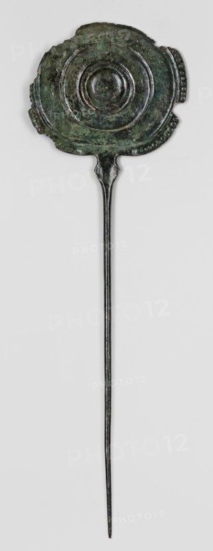 Pin, unknown Luristan workshop. A long pin terminating in a head in the form of an oval shield with geometric decoration. The shaft, circular in cross-section, thickens and widens rhomboidally at the top to form a connector to the shield, which is decorated on one side with punching and engraving. Ornamentation on the shield is arranged concentrically: central round knob with two intersecting engraved bands filled with fine dots; three convex concentric stripes central stripe incised; interwoven braids with dotted interiors in the spaces between stripes; a row of larger dots between the first stripe and the boss; the edge band decorated with a single row of convex dots. The decorative sequence is interrupted at the lower part of the shield where it connects to the pin: on a short section of the edge band the convex dots are replaced by dots, and diagonal incisions appear on the third strip. Engraved geometric ornament also appears on the upper part of the rhomboid connector. Basketry o...