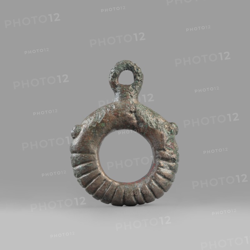 Decorative pendant from a horse harness, unknown Near Eastern workshop. The pendant is in the form of a thick ring with a round eyelet at the top. The lower half of the ring is decorated with 16 notches. On both sides, the notching terminates in a pair of small round knobs. The pendant was probably an element of a harness. 9th–6th century BC end of the 2nd millennium–first half of the 1st millennium BC; Iron Period. Luristan, Near East. Harness component, pendant; material culture; decoration. Weight 94 g; diameter 5.5 cm; thickness 1.2 cm., A circular metal ring with a rough textured surface and a small loop at the top, showing a patina of green and brown with hints of darker gray and reddish tones, the lower half of the ring features a series of carved ridges creating a fluted pattern, the object rests on a smooth light gray background and casts a soft shadow beneath it.