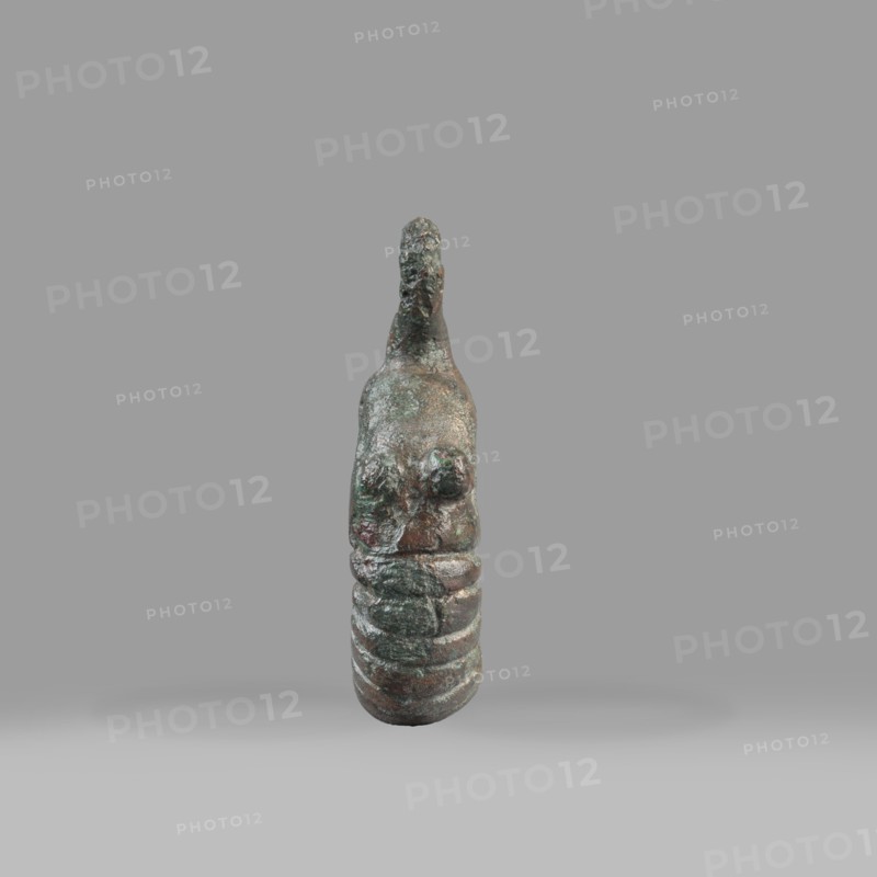 Decorative pendant from a horse harness, unknown Near Eastern workshop. The pendant is in the form of a thick ring with a round eyelet at the top. The lower half of the ring is decorated with 16 notches; on both sides the notching terminates in a pair of small round knobs. The pendant was probably an element of a harness. 9th–6th century BC end of the 2nd millennium–first half of the 1st millennium BC; Iron Period. Provenance: Near East; origin: Luristan. Materials: metal; harness component/pendant; decoration. Weight: 94 g; diameter: 5.5 cm; thickness: 1.2 cm., A small sculptural object stands upright against a plain gray background, showing a textured, weathered surface with a mix of green, brown, and dark bronze tones; the form tapers toward the top with an irregular protrusion, a rounded upper section with two raised rounded areas on the front, and a segmented lower portion with a series of rounded horizontal ridges; lighting casts a soft shadow beneath the object.