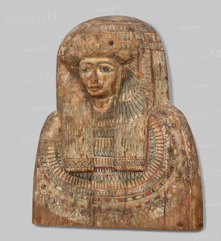 Fragment of a sarcophagus lid. Part of the lid of an anthropoid woman's sarcophagus. Large, striped wig falling on the breasts at the front, decorated with four diadems of lotus petals and vulture wings, and bands of rosettes at the ends. On the breast is a multi-row necklace, with lotus flowers and rosettes forming the lower row. Lotuses, vulture vulture wings, element of the so-called vulture diadem, necklace, wigs, funerary equipment, lotus flower decorative motif. Egyptian art. c. 945–c. 735 BC; Third Intermediate Period, 22nd Dynasty, Egypt. Sarcophagus; height 62 cm, width 49 cm, depth 20 cm. /author., A painted wooden funerary panel showing the upper torso and head of a figure with a stylized wig or headdress, carved with a rounded top and flat bottom. The surface shows worn paint and areas of chipped wood. The face has almond-shaped eyes, a defined nose, and a small mouth, all rendered in muted tones. The wig or headdress falls to shoulder level with linear carved sections and ...