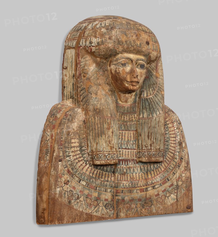 Fragment of a sarcophagus lid. Part of the lid of an anthropoid woman's sarcophagus. Large, striped wig falling on the breasts at the front, decorated with four diadems of lotus petals and vulture wings, and bands of rosettes at the ends. On the breast is a multi-row necklace, with lotus flowers and rosettes forming the lower row. Lotuses, vulture vulture wings, element of the so-called vulture diadem, necklace, wigs, funerary equipment, lotus flower decorative motif. Egyptian art. c. 945–c. 735 BC; Third Intermediate Period, 22nd Dynasty, Egypt. Sarcophagus. Height 62 cm; width 49 cm; depth 20 cm., A carved wooden funerary panel shaped like a bust with a stylized head and shoulders, showing worn painted surface and areas of flaking and loss; the face has carved eyes, nose, and mouth with traces of darker pigment around the eyes and mouth, and the hair or headdress is rendered as a rounded, layered wig that extends down over the shoulders. The chest area is decorated with a wide, ornat...