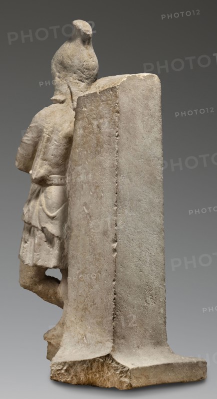 Fragment of a monopodium, unknown Roman workshop, Roman sculpture of Attys myth., 2nd century, Roman Empire 27 BC–476, sculpture; height 77 cm, width 31 cm, depth 34 cm, weight 64 kg., A pale stone sculpture viewed from an angle showing the back and side of a standing figure partially obscured by a tall rectangular slab; the figure wears a short garment with folds and a belt and has one leg bent with a foot resting on a base. The stone surfaces show rough and smoother textures with weathered edges and tool marks. Visible colors include beige, off-white, light gray, tan, and hints of brown.