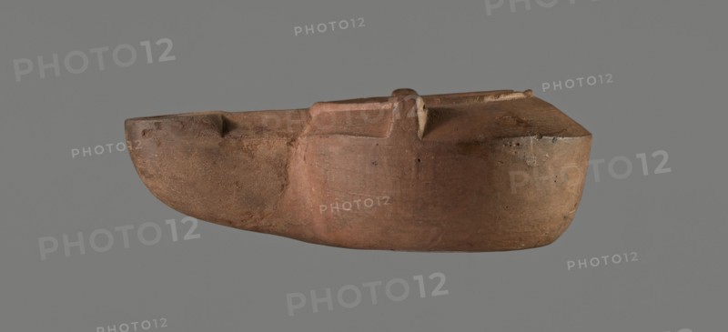 Lamp with a tragic mask, unknown Italian workshop. Oil lamp, single-burner, Roman, Buchi type IX-b = Loeschcke type IXb, probably 2nd half 1st–beginning 2nd century. From a mold. Round vessel with arms drooping to the outer, separated from the disc by a convex edge; two bars arranged symmetrically on the arms. Concave, flat disc with two olive holes located on either side of a convex theatrical tragic mask. Medium-length wick channel decorated with a central groove and a small ventilation hole in the groove; rounded end of the burner. Slight trace of use dark discoloration near the wick hole. On the bottom, a convex inscription workshop trademark: COMMUNIS. Height c. 9 cm, width c. 4.4 cm, depth c. 3.3 cm., A single elongated object resembling a carved vessel or model boat fills the center of the image, shown in profile against a plain gray background. The object has an overall earthy brown and reddish-brown surface with areas of darker brown and muted orange tones, and a few lighter b...