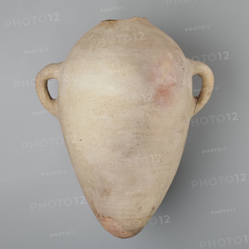 Large ovoid amphora with rounded foot, clay MD marl, Vienna System. Body wheel-made; hand-made foot. Two small vertical handles placed at greatest bulge at base of the sloping arms; sloping arms symmetrically placed with two black-painted inscriptions labels/dockets on their bases. Above one handle, slightly toward the center, are impressed cartouches of Hatshepsut and Thutmose III and a snake hieroglyph dz. Potter’s mark of two intersecting lines beneath one handle. Surface even; color very light brown. Incomplete—neck and spout missing. New Kingdom, 18th Dynasty reign of Hatshepsut and Thutmose III, ca. 1550–1295 BC. Preserved height 49.5 cm; diameter at belly 30.5 cm., A large, rounded clay vessel with a tapered pointed base and a narrow top, shown against a gray background; the vessel has a matte, textured surface in pale beige and tan tones with areas of light pink and faint reddish patches, and it features a pair of looped handles on opposite sides that are similar in color and t...