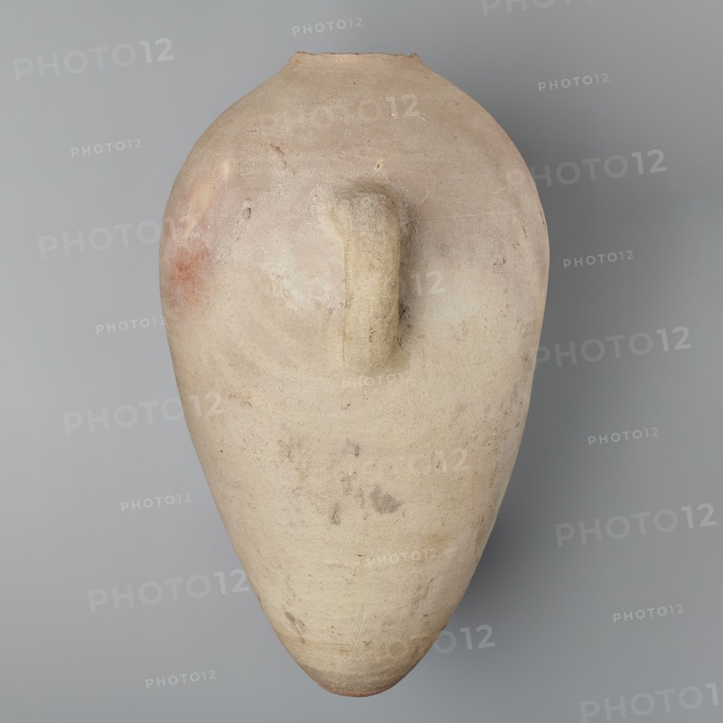 Large ovoid amphora with rounded foot, clay MD-type marl, Vienna System; body wheel-made, foot hand-made; surface even; color very light brown. Two small vertical handles placed at widest point near base of the sloping neck; neck and spout missing. Sloping handles symmetrically painted with black ink inscriptions labels/dockets. Above one handle, slightly toward center, are impressed cartouches of Hatshepsut and Thutmose III and a snake hieroglyph dz. Under one handle a potter’s mark of two intersecting lines. New Kingdom, 18th Dynasty reign of Hatshepsut and Thutmose III, ca. 1550–1295 BC; provenance: Deir el-Medina grave 1370 f. Preserved height 49.5 cm; belly diameter 30.5 cm. Analogies: amphora from the tomb of Hatnefer and Ramose with hieratic inscription dating to year 7 of the joint reign; R. Holthoer, New Kingdom pharaonic sites., A large, elongated clay vessel with a rounded top and tapered bottom is shown against a smooth background. The vessel has a single short handle attac...