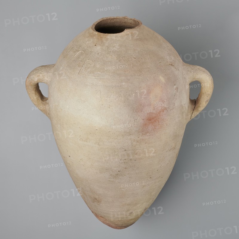 Large amphora with an ovoid shape and a rounded foot. Two small vertical vices placed on the body at the point of its greatest bulge at the base of the arms. The foot is hand-made; the body was wheel-made. Made of MD type marl clay Vienna System. Surface even. Sloping arms, with inscriptions in black paint labels, dockets symmetrically placed on both sides of the body. Above one of the vices, slightly toward the center of the body, are impressed cartouches of Hatshepsut and Thutmose III and a snake hieroglyph dz. Under one of the vices, a potter's mark of two intersecting lines. Very light brown color. Incomplete — neck and spout missing. New Kingdom, 18th Dynasty reign of Hatshepsut and Thutmose III, ca. 1550–1295 BC. Two cartouches of Hatshepsut and Thutmose III; two labels on the body. Preserved height 49.5 cm; belly diameter 30.5 cm., A large, rounded clay vessel with a tapering pointed base and a wide, irregular mouth, shown against a flat neutral background. The vessel has a pair...