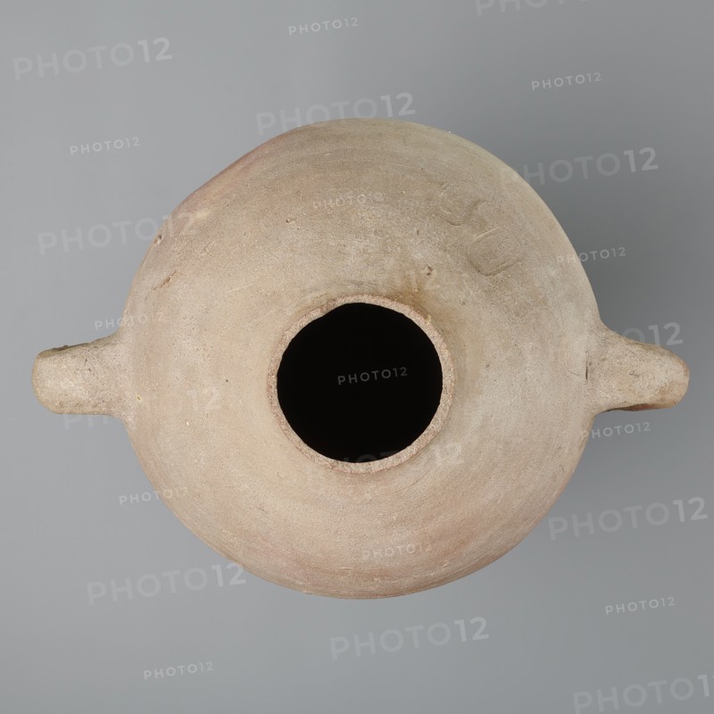 Large amphora with an ovoid shape and a rounded foot. Two small vertical vices placed on the body at the point of its greatest bulge at the base of the arms. The foot is hand-made, the body wheel-made. Made of MD type marl clay Vienna System. Surface even. Sloping arms, along their bases on both sides of the body, are symmetrically placed with inscriptions in black paint labels, dockets. Above one of the vices, slightly toward the center of the body, are impressed cartouches of Hatshepsut and Thutmose III and a snake hieroglyph dz. Under one of the vices, a potter's mark of two intersecting lines. Color very light brown. Vessel incomplete — neck and spout missing. Cartouches of Hatshepsut and Thutmose III; two labels on the body. New Kingdom, 18th Dynasty reign of Hatshepsut and Thutmose III, ca. 1550–1295 BC; findspot: Deir el-Medina grave 1370 f. Preserved height 49.5 cm; belly diameter 30.5 cm., A top-down view of a rounded vessel with a wide central opening and a few small protrudi...