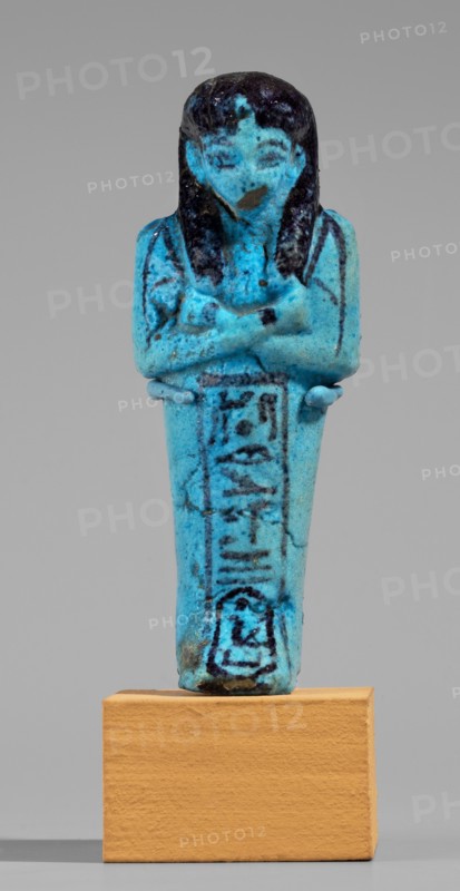 Queen Maatkare's shabti, Divine Wife of Amun, cartouches, hieroglyphs, uraeus iconographic, Maatkare ca. 1069–945 BC, wigs, funerary figurines, grave goods, mummy-shaped figurine. Arms crossed on the chest, right folded over left. Hoes in each hand. On the back is a basket slung on strings over both shoulders. Facial details, wig, and tools painted in a dark color. Three-part smooth wig. Uraeus above the forehead. Bracelets on the wrists. Painted inscription on the front of the legs in one wide column framed with lines: Shd Osiris, the wife of the god, the lord of both lands, Maat-ka-re. Third Intermediate Period; 21st Dynasty, Egypt. Shabti, height 12 cm, width 4.4 cm., A small blue figurine of a standing human figure with crossed arms, mounted on a rectangular tan base, shown against a plain light gray background. The figure is predominantly bright turquoise blue with darker blue to black coloration on the hair, shoulders, and facial features, and some darker blue markings on the tor...