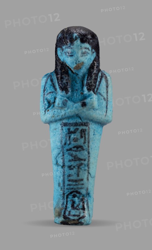 Queen Maatkare's shabti, Divine Wife of Amun, cartouches, hieroglyphs, uraeus iconographic, Maatkare ca. 1069–945 BC, wigs, funerary figurines, grave goods, mummy-shaped figurine. Arms crossed on the chest, the right folded over the left, holding hoes in each hand. On the back is a basket slung on strings over both shoulders. Facial details, wig, and tools painted in a dark color. On the head is a three-part smooth wig with a uraeus above the forehead. Bracelets on the wrists. Painted inscription on the front of the legs in one wide column framed with lines reading: Shd Osiris, the wife of the god, the lord of both lands, Maat-ka-re. Third Intermediate Period, 21st Dynasty, Egypt; shabti, height 12 cm, width 4.4 cm., A small figurine in shades of bright turquoise and blue stands upright against a gray background, depicting a human form with folded arms and a long, tapered lower body; the head and hair are darker, nearly black, with visible streaks and textured surface, and there are da...