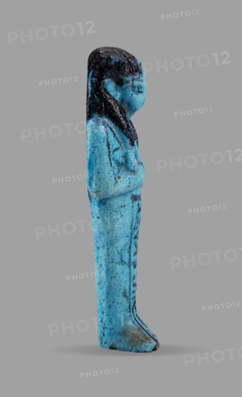 Queen Maatkare's shabti, Divine Wife of Amun, cartouches, hieroglyphs, uraeus iconographic, Maatkare ca. 1069–945 BC, wigs, funerary figurines, grave goods, mummy-shaped figurine. Arms crossed on the chest, the right folded over the left. Hoes in each hand. On the back is a basket slung on strings over both shoulders. Facial details, wig, and tools painted in a dark color. On the head is a three-part smooth wig with a uraeus above the forehead. Bracelets on the wrists. Painted inscription on the front of the legs in one wide column framed with lines: Shd Osiris, the wife of the god, the lord of both lands, Maat-ka-re. Owner: Osiris, the divine wife of the lord of both lands, Maatkare; ca. 1069–ca. 945 BC; Third Intermediate Period; 21st Dynasty, Egypt. Shabti; height 12 cm, width 4.4 cm., A small standing figurine shown in profile with a smooth elongated body and a rounded head, displaying a few raised arms folded across the chest and a long hairstyle that falls along the back; the sur...